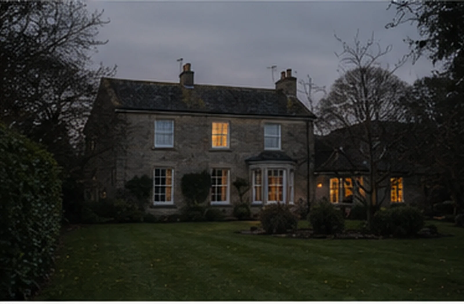 A well-kept house at dusk seen from the garden, one lamp lit inside