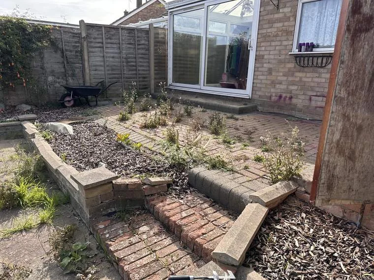 Block paving beside a bungalow with weeds through the joints and the bark border grown over